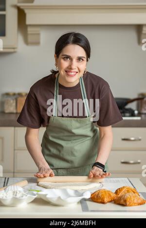 Mignon jeune brunette femme souriante dans un tablier de cuisine est la cuisson maison de la cuisson dans la cuisine. Banque D'Images