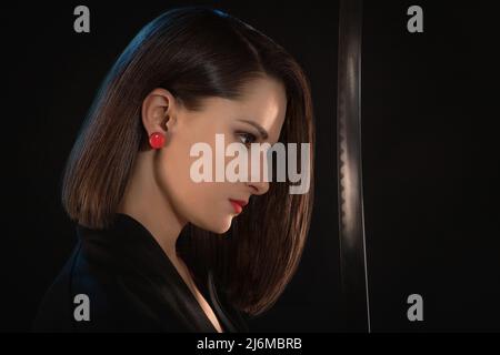 Portrait d'une jeune femme avec des lèvres rouges et des boucles d'oreilles devant une lame aiguisée sur un fond noir. Banque D'Images