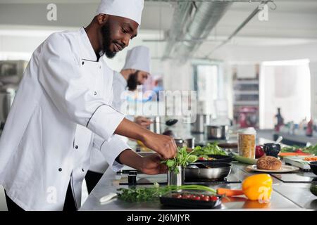 Chef cuisinier afro-américain professionnel cueillant des herbes vertes fraîches pour améliorer le goût du plat gastronomique. Un cuisinier masculin portant un uniforme de cuisine préparant des repas biologiques dans la cuisine du restaurant. Banque D'Images