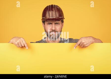 homme mature travailleur dans le casque pointant le doigt sur le papier jaune pour l'espace de copie, annonce Banque D'Images