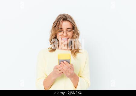 Une jeune femme blonde souriante et satisfaite porte des lunettes utilise son smartphone tout en se tenant sur un fond blanc Uni Banque D'Images