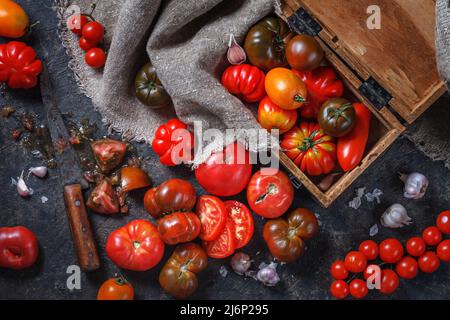 Des tomates mûres fraîches et colorées de différentes variétés de limes, des tranches de tomates, de l'ail et un couteau vintage se trouvent sur une table sombre. Les légumes biologiques collectent Banque D'Images