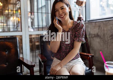 Femme asiatique souriante parlant au téléphone portable dans Cafe, gérer son horaire d'affaires, planifier des réunions, parler avec un client ou des amis, assis à la table Banque D'Images