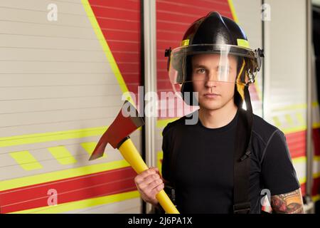 Photo d'un pompier portant un casque avec une hache contre un moteur d'incendie. Banque D'Images