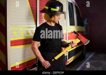 Photo d'un pompier portant un casque avec une hache contre un moteur d'incendie. Banque D'Images