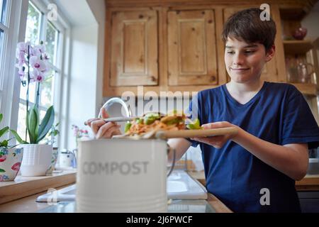 Garçon dans la cuisine faisant du compost raclant les restes de légumes dans le bac Banque D'Images