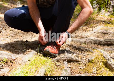 un adulte, un homme qui se croise dans un pantalon bleu et un gros plan de ses mains, enfile les lacets de ses baskets rouges sur quelques racines dans une forêt, après avoir marché Banque D'Images