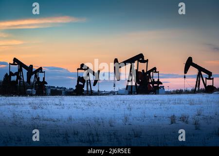 Vérins à pompe à gaz et à pétrole travaillant sur un champ couvert de neige au coucher du soleil dans le comté de Rocky View Alberta Canada. Banque D'Images