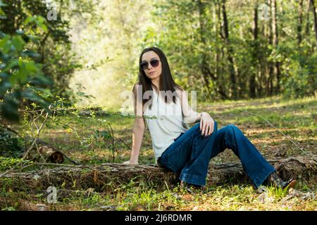 Une belle jeune fille sérieuse brune portant des lunettes de soleil regardant loin de l'appareil photo vers l'avenir dans une zone boisée au printemps Banque D'Images