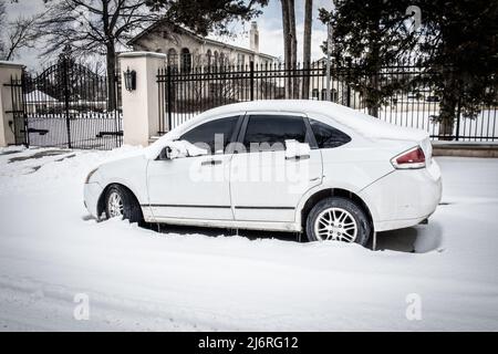 Voiture blanche sale avec des éraflures garées dans la neige profonde sur la rue résidentielle à l'extérieur de la grande clôture avec manoir et domaine de luxe en arrière-plan. Banque D'Images