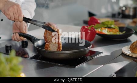 Chef cuisinier professionnel cuisant le steak de porc dans une poêle, à l'aide d'huile de tournesol biologique et d'ustensiles de cuisine pour préparer de la viande fumée. Cuisinez un délicieux plat gastronomique. Gros plan. Prise de vue à main levée. Banque D'Images