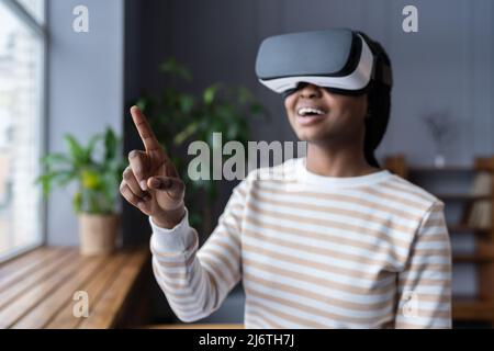 Fille noire en lunettes de réalité virtuelle se sentant excitée et stupéfait, touchant l'objet dans le monde virtuel Banque D'Images