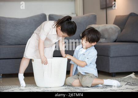 Par le canapé du salon, une petite fille est prête à s'asseoir dans le panier à vêtements et le petit garçon l'aide - photo de stock Banque D'Images