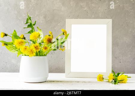 Création d'une maquette. Cadre blanc sur table en bois blanc avec fond en béton gris et pissenlits dans un vase Banque D'Images