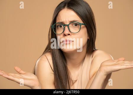 Perplexe la jeune femme adulte avec les bras sortis, haussant les épaules, en disant: Qui se soucie, donc quoi, Je ne sais pas. Studio isolé tourné sur fond beige. Banque D'Images