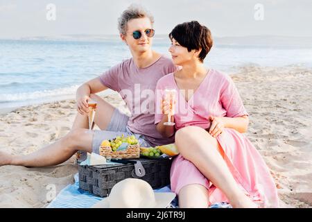 Couple ayant un pique-nique à la plage de bord de mer. Homme et femme amoureux tenant des verres avec du vin ou du champagne, célébrant le moment avec fond de mer. Banque D'Images