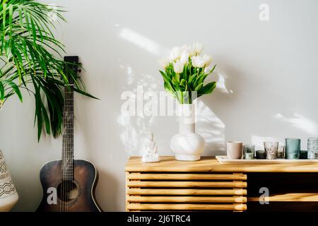 Décoration intérieure minimaliste scandinave - bouquet de fleurs de tulipe blanches dans un vase, bougies, statuette de Bouddha sur une armoire en bois. Plante de maison verte Banque D'Images