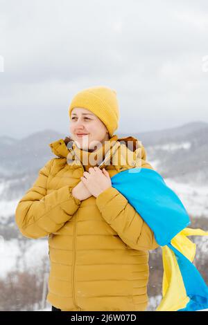 Jeune fille portant un drapeau national bleu et jaune de l'Ukraine sur un fond de montagnes carpathes enneigées et de ciel bleu. Femme en veste orange et chapeau jaune. Jour de la Constitution. Banque D'Images