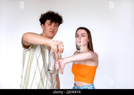 Portrait d'un jeune couple sérieux posant sur fond blanc. Jeune homme bouclés confiant, femme montrant le signe du pouce vers le bas. Banque D'Images