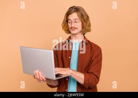 Portrait d'un beau gars à cheveux rouges, doué et branché, utilisant un ordinateur portable écrivant un e-mail isolé sur fond beige pastel Banque D'Images