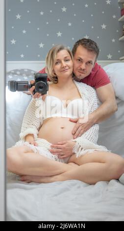 Couple enceinte prenant le selfie dans le miroir avec un appareil photo numérique. Mari embrasse sa femme. Famille - homme et femme. Grossesse, maternité concept. Banque D'Images