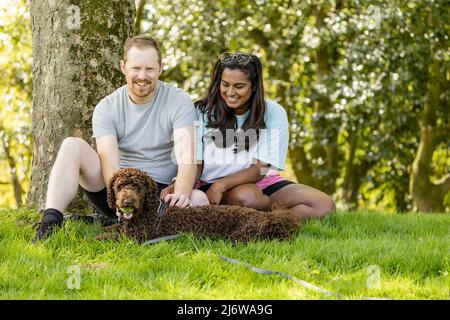 Jeune homme blanc et sa petite amie asiatique assis sous un arbre avec leur chiot de coolé de couleur chocolat Banque D'Images