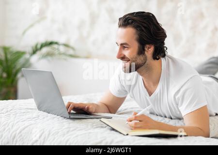 jeune homme positif, posé sur le lit, utilisant un ordinateur portable, prenant des notes Banque D'Images