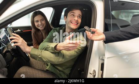 Heureux couple millénaire prenant la clé de voiture du vendeur d'auto, assis à l'intérieur de l'automobile moderne à la concession, panorama Banque D'Images