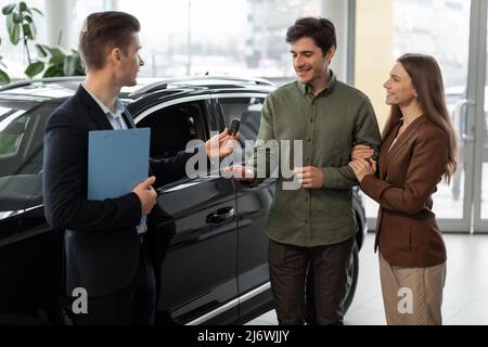 Concept d'achat ou de location de voiture. Jeune couple joyeux prenant la clé de voiture du vendeur d'auto à la concession moderne Banque D'Images