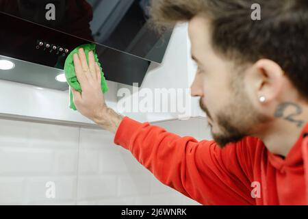 Jeune homme portant une hotte aspirante rouge ou un ventilateur de cuisine avec un chiffon et un détergent dans la cuisine. Banque D'Images