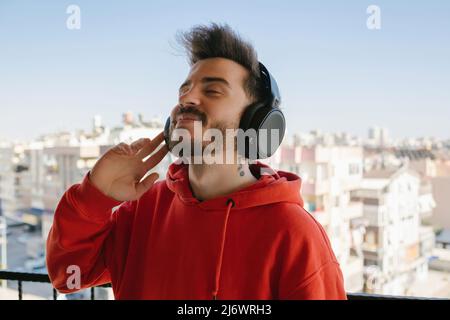 Jeune homme heureux portant un sweat à capuche rouge écoutant de la musique avec un casque. Profiter de sa musique préférée. Ecoute et sourit en restant debout Banque D'Images