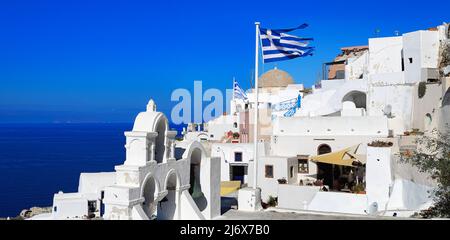 Oia, Santorini, Iles grecques, Grèce - églises blanchies à la chaux, dôme et clocher, drapeau grec et maisons, perchées sur une falaise sous un soleil éclatant Banque D'Images