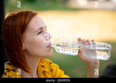 Une femme d'âge moyen à tête rouge boit de l'eau dans une bouteille. Étancher la soif, maintenir l'équilibre de l'eau lors d'une journée chaude. Banque D'Images