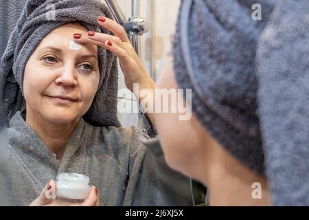 Une jeune femme souriante dans une serviette applique de la crème pour le visage. Le concept de rajeunissement de la peau. Le concept de soins de la peau Banque D'Images