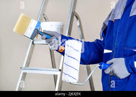 Le constructeur est un peintre en costume bleu, un pinceau dans sa main et des seaux avec des produits pour la restauration et la peinture du mur, sur le constructi Banque D'Images