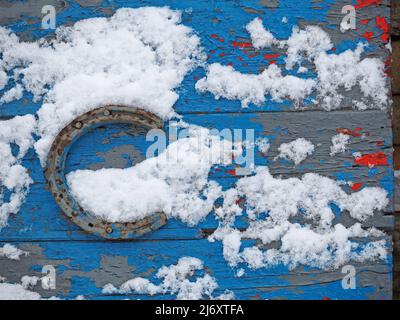 neige fraîche nichée sur le fer à cheval rouillé clouée à la porte de ferme de bois détachée avec couche de finition bleue écaillée sur des couches de peinture contrastantes gris et rouge Banque D'Images