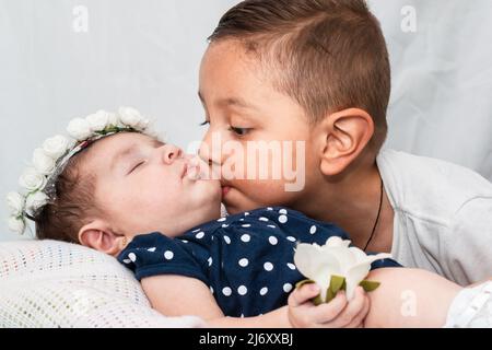 petit garçon latino embrassant la joue de sa petite sœur, le bébé est allongé sur un oreiller blanc, avec ses yeux fermés, une couronne de roses blanches et tenant un r Banque D'Images