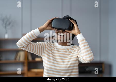 Stupéfait femme afro-américaine testant un casque VR moderne de haute technologie, obtenant une expérience 3D à la maison Banque D'Images