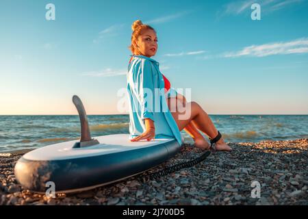 Une belle femme caucasienne plentue pose assis sur un plateau de sup. Vue arrière. En arrière-plan, le ciel est l'océan. Le concept de loisirs sportifs b Banque D'Images