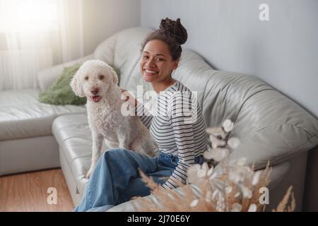 Sourit une jolie femme de course mixte assise dans une pièce confortable sur un canapé avec un chien de chien en forme de coolé blanc Banque D'Images