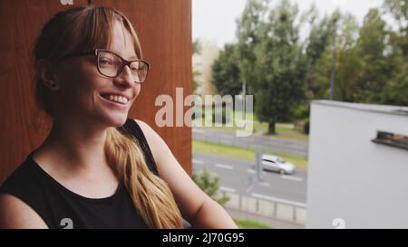 Gros plan portrait extérieur d'une femme caucasienne aux cheveux rouges debout sur son balcon, regardant la circulation, souriant. Photo de haute qualité Banque D'Images