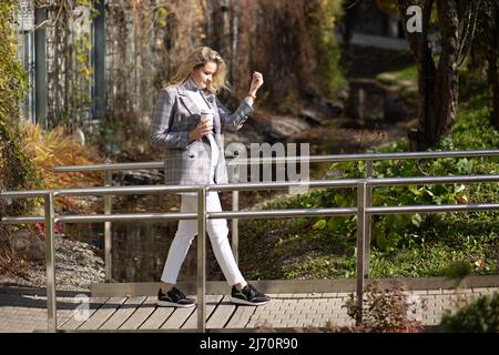 Femme élégante appréciant aller le long du pont à travers le ruisseau avec café en automne vue sur le parc de la ville. Une femme d'affaires qui marche et se détend dans la rue Banque D'Images