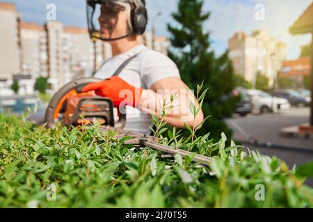 Jardinier masculin compétent en uniforme, masque de sécurité et gants coupant des buissons pendant l'été. Homme caucasien utilisant un couteau à haie à essence tout en prenant soin des plantes vertes à l'air frais. Banque D'Images