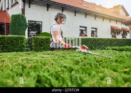 Homme caucasien en uniforme, lunettes de protection et masque de taille des bagues surcultivées avec coupe électrique. Jardinier compétent prenant soin des plantes sur l'arrière-cour pendant l'été. Banque D'Images