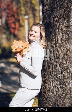 La femme heureuse se tient debout contre le tronc d'arbre avec des feuilles mortes dans le parc de ville d'automne le jour ensoleillé. Une femme adulte tient le feuillage jaune entre les mains et sourit Banque D'Images