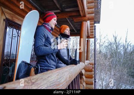 Un gars dans un chapeau d'orange en hiver boit le thé d'un thermos pendant des vacances d'hiver dans les montagnes. Il se tient sur le balcon d'une maison en bois. Port Banque D'Images