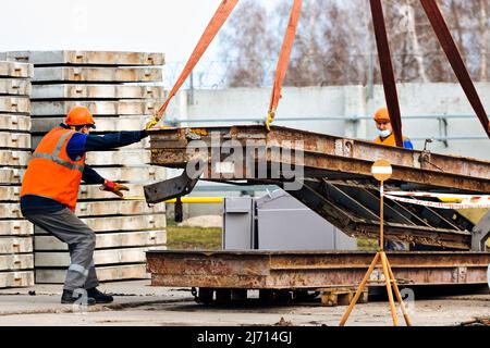 L'aiguillon dans le casque et le gilet contrôle le déchargement des structures métalliques sur le chantier de construction. Le handyman blanc décharge la charge. Banque D'Images