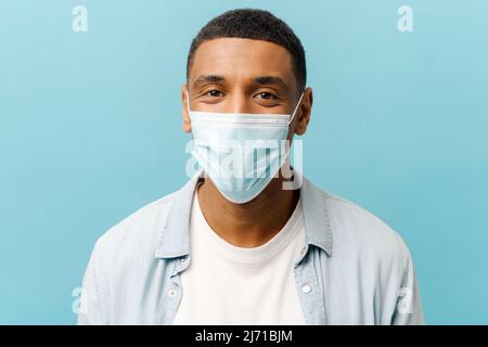 Jeune homme afro-américain positif avec coupe courte portant un masque médical de protection regardant la caméra isolée sur fond bleu, homme se protégeant contre les maladies virales pendant la pandémie Banque D'Images