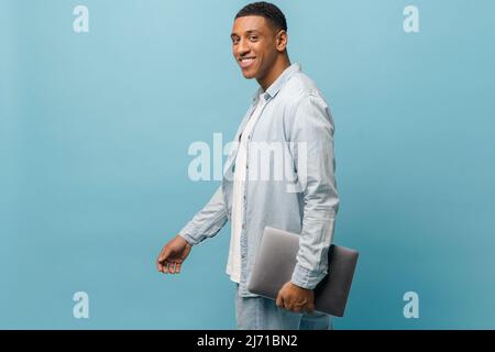 Sympathique jeune afro-américain étudiant dans une chemise décontractée portant un ordinateur portable isolé sur fond bleu, souriant multiracial indépendant homme regardant la caméra Banque D'Images