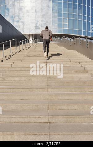 Vue en contre-plongée d'un athlète masculin noir anonyme en train de courir des marches en béton vers un bâtiment de bureau contemporain tout en ayant l'entraînement en plein air Banque D'Images